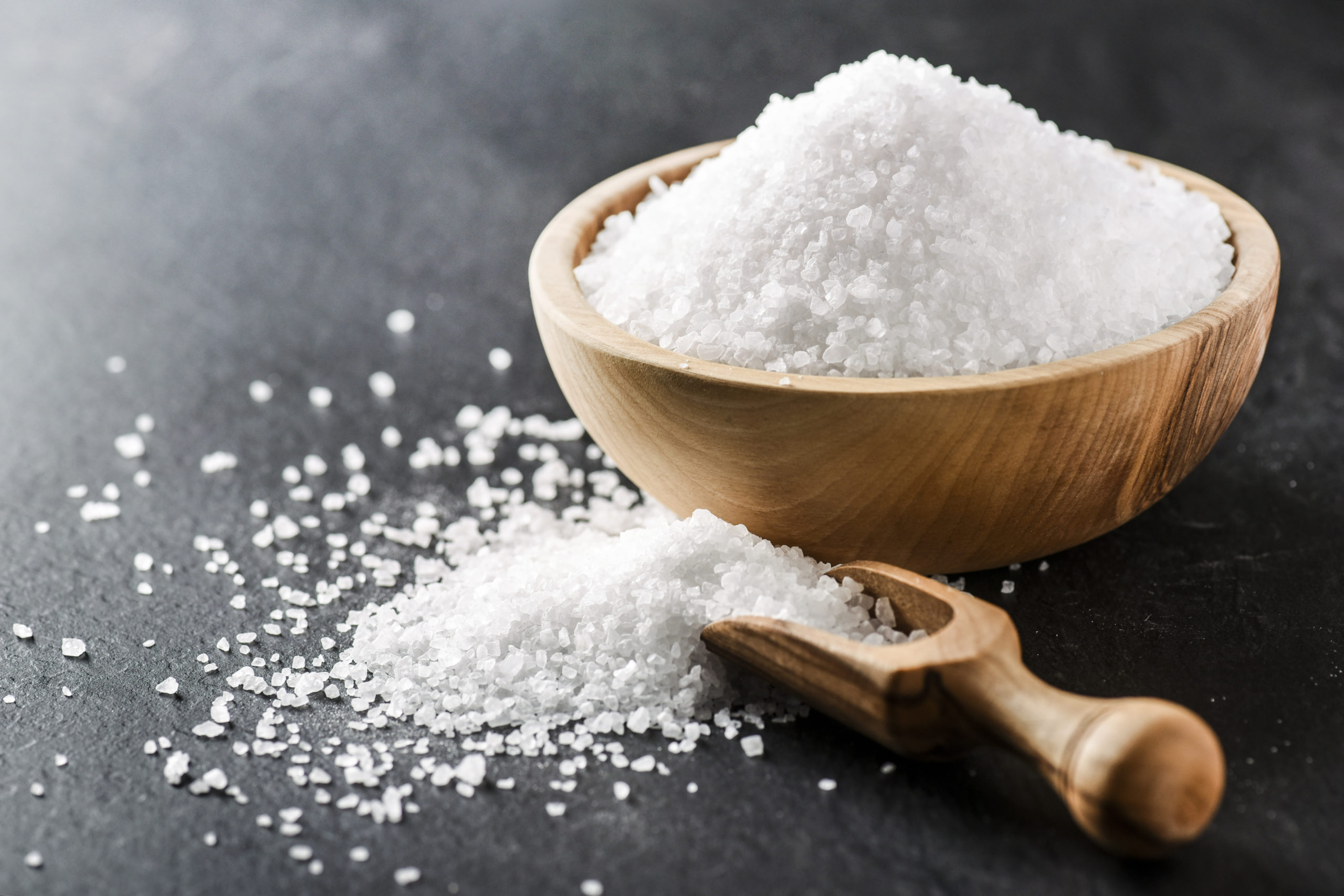 Salt in wooden olive bowl with scoop on stone table. Salt top view. Sea salt on black background.